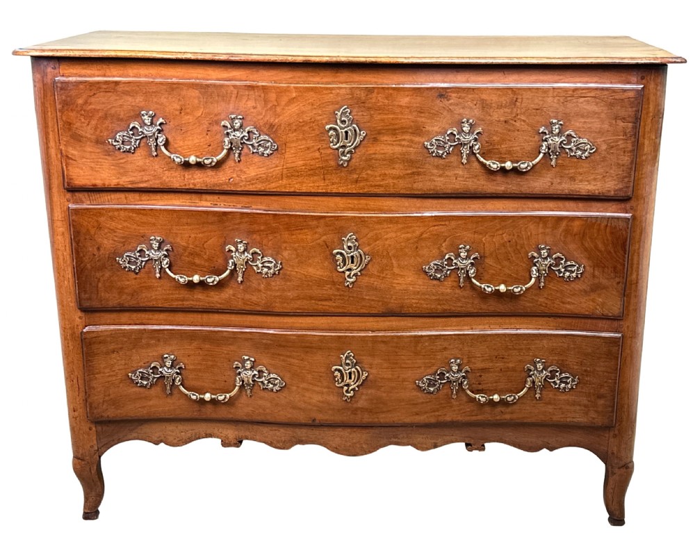 An 18th-century French provincial walnut commode chest featuring three full-width drawers decorated with elaborate brass handles and keyhole escutcheons, set above a scalloped apron and short cabriole legs. View XV
