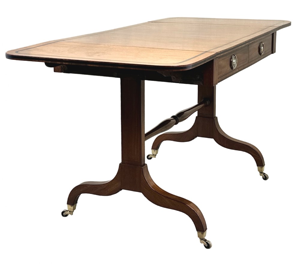 A 19th-century Regency mahogany sofa table featuring two drop leaves, two frieze drawers with octagonal brass handles, a turned stretcher, and splay legs on brass castors. View IV