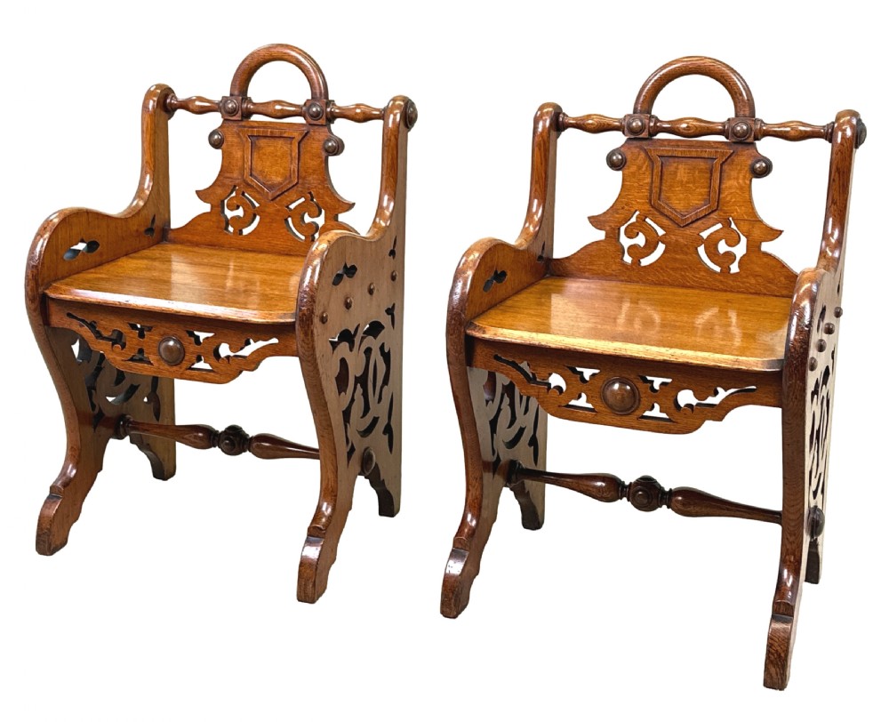 A matched pair of 19th-century carved oak hall seats featuring arched top handle rails, shield-carved back panels, pierced side aprons, and turned cross stretchers.