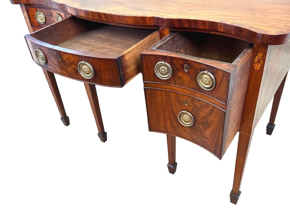 A small Georgian mahogany sideboard featuring a serpentine front, floral marquetry inlay, multiple drawers with brass ring handles, and tapered square legs with spade feet. View VI