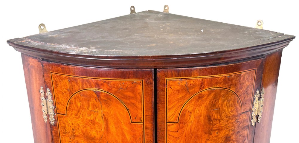 A bow-fronted Georgian corner cupboard featuring highly figured burr walnut veneers, inlaid crossbanding, double arched panelled doors, and shaped brass hinges. View VI