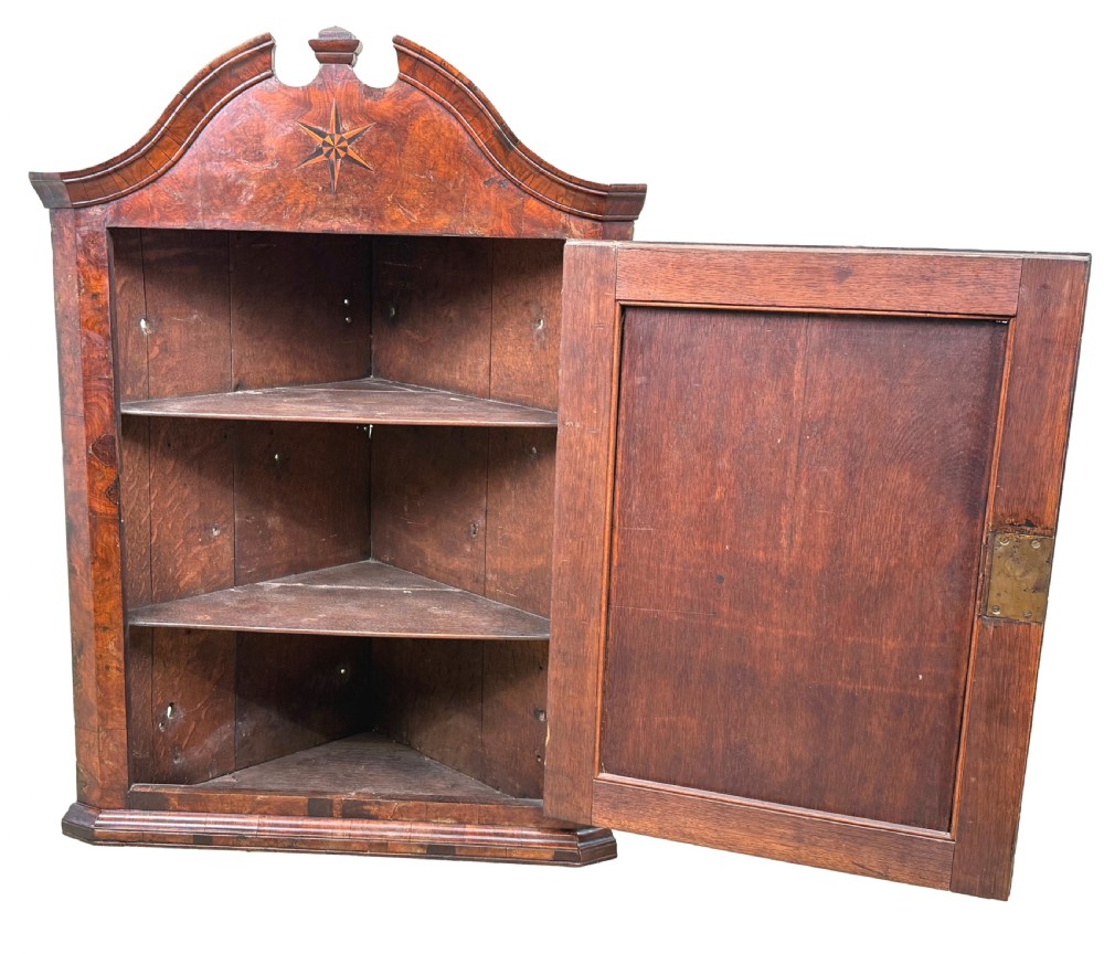 An antique Queen Anne burr walnut wall-hanging corner cupboard featuring an arched pediment with an inlaid star motif and a single fielded panel door. View IV