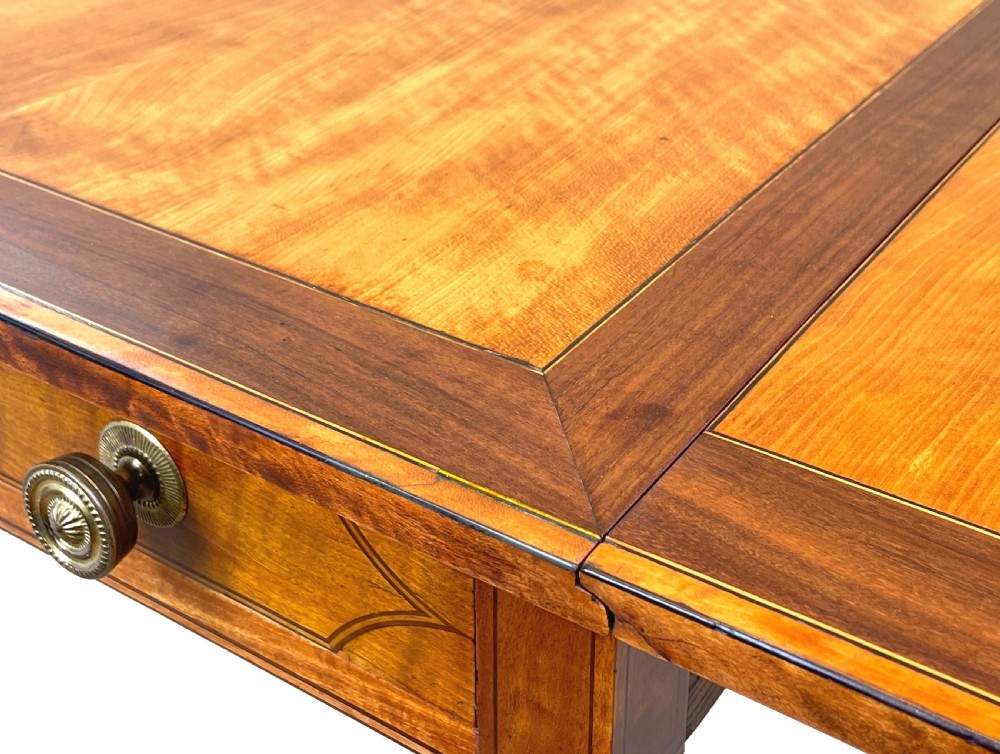 A late 18th-century Georgian satinwood Pembroke table featuring two hinged drop leaves, dark wood crossbanding, a drawer with brass knobs, and tapered square legs with castors. View VIII
