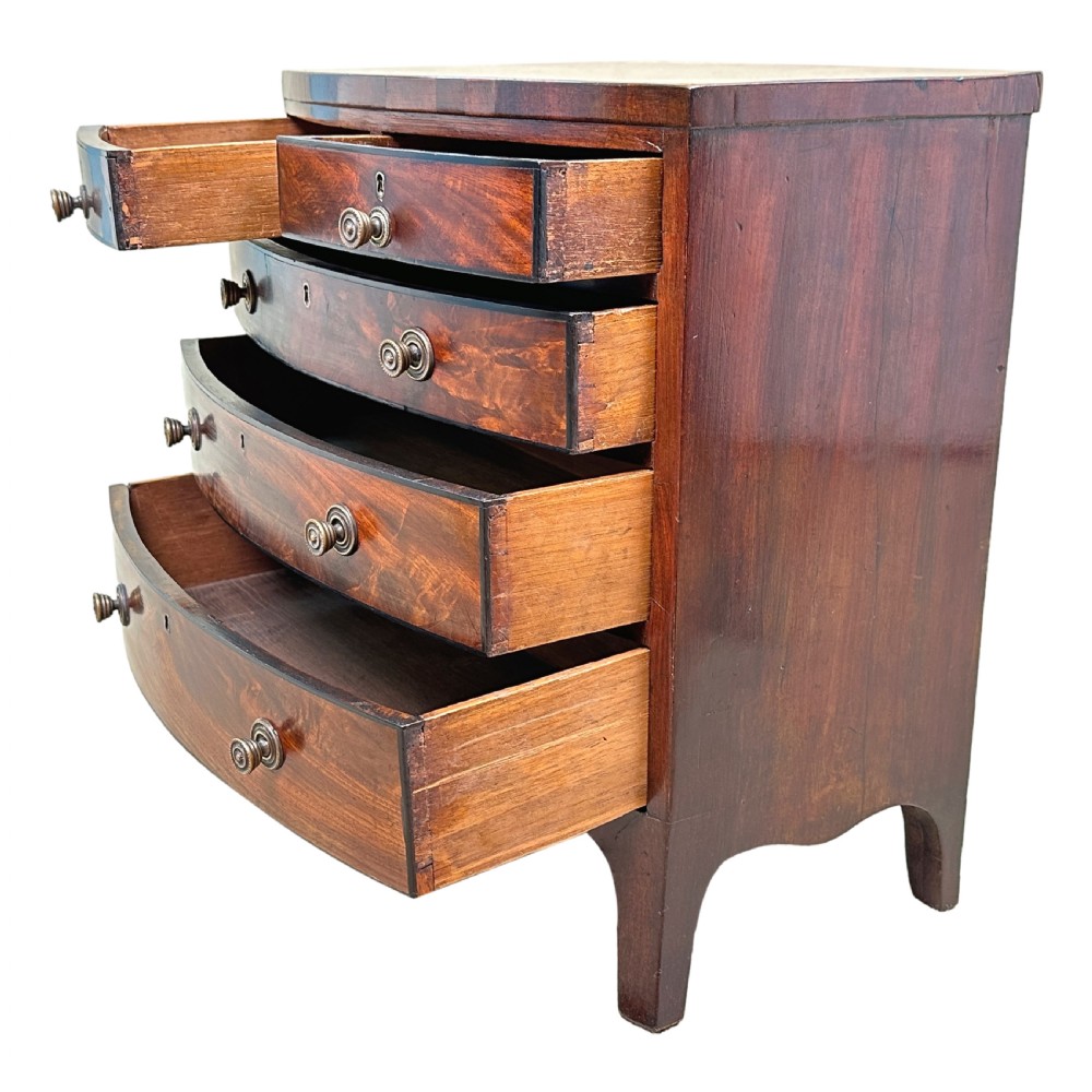 A Georgian mahogany miniature bowfront chest of drawers with flame mahogany drawer fronts, turned brass handles, and splayed bracket feet. View IV