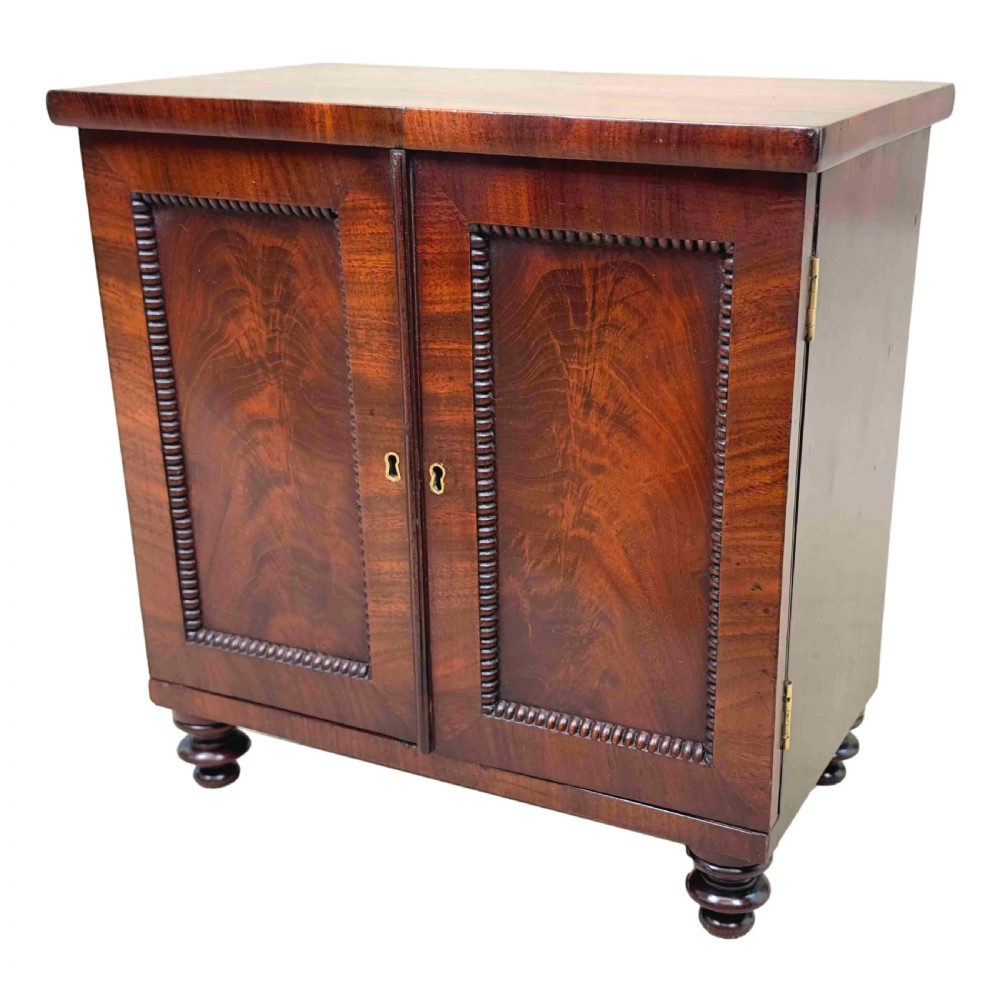 A Regency flame mahogany twin-door miniature table cabinet with turned feet, bead moulding, and brass escutcheons.