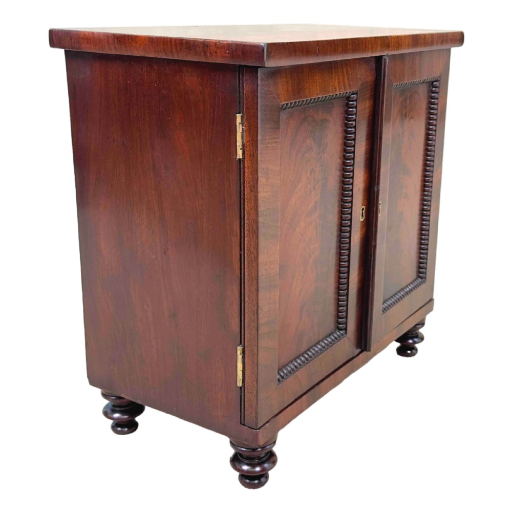 A Regency flame mahogany twin-door miniature table cabinet with turned feet, bead moulding, and brass escutcheons. View VII