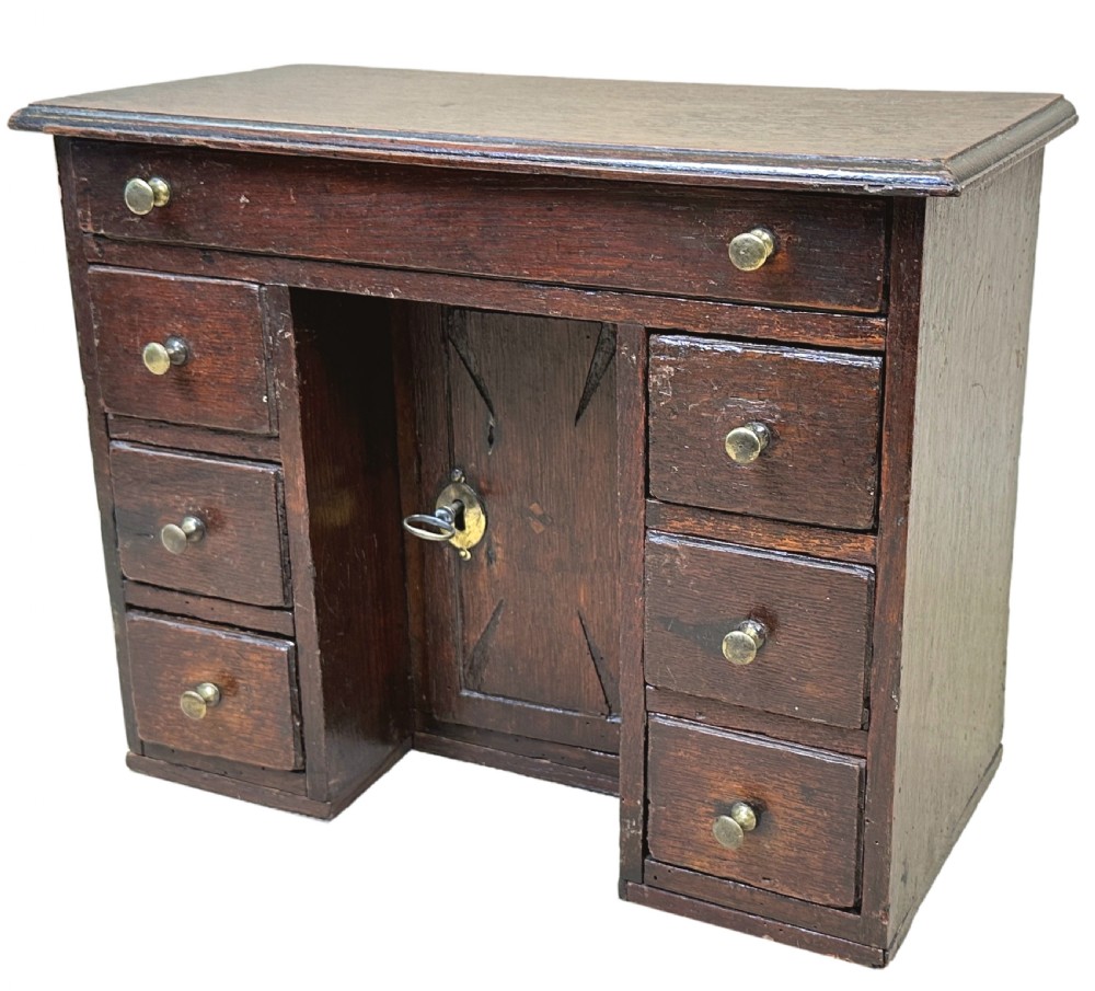 A dark oak miniature kneehole desk featuring a single top drawer over six side drawers and a central cupboard with a key and brass knobs. View III