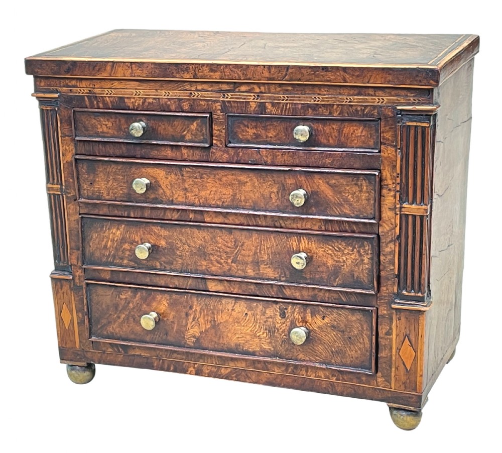 A Regency era miniature chest of drawers crafted from burr oak with geometric inlay, brass knobs, reeded pilasters, and small turned ball feet. View II