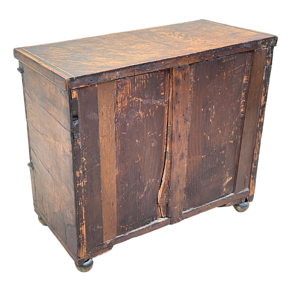 A Regency era miniature chest of drawers crafted from burr oak with geometric inlay, brass knobs, reeded pilasters, and small turned ball feet. View VIII