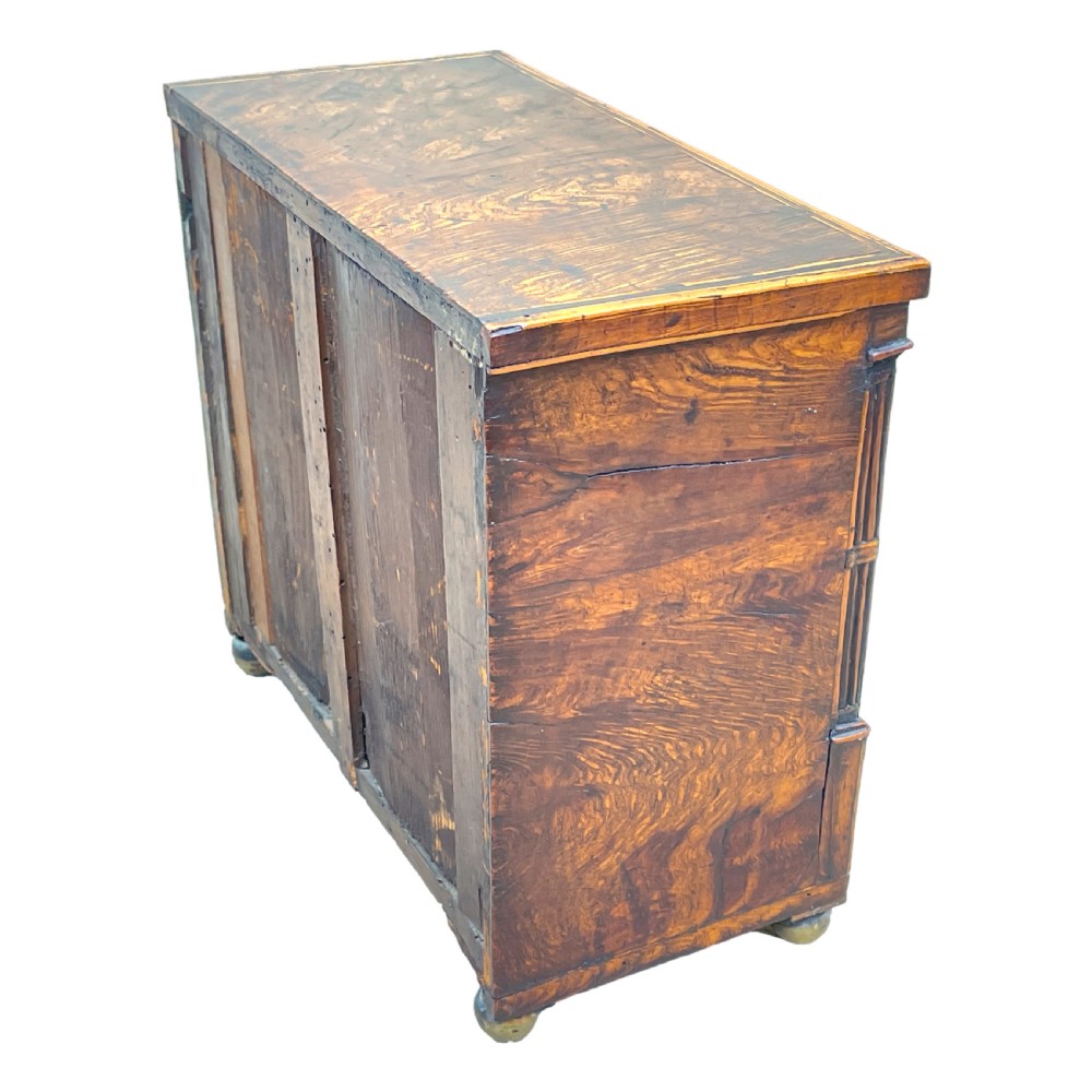 A Regency era miniature chest of drawers crafted from burr oak with geometric inlay, brass knobs, reeded pilasters, and small turned ball feet. View IX