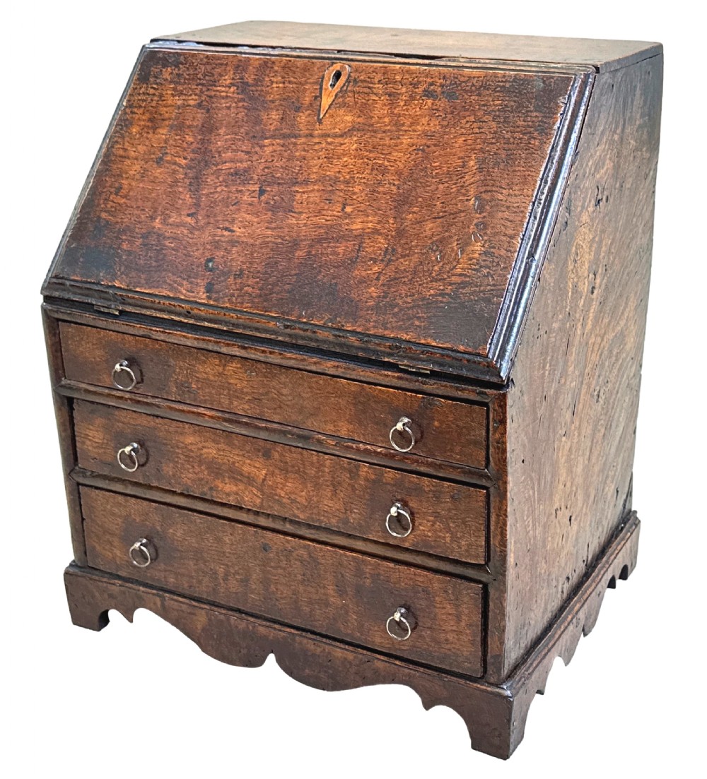 An 18th-century Georgian tabletop bureau made of dark warm elm wood, featuring a sloped writing flap above three drawers with brass ring pulls and a scalloped base.
