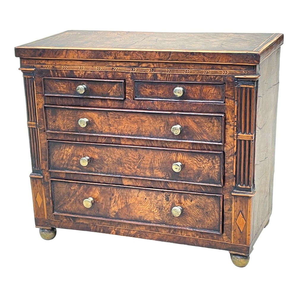 A Regency era miniature chest of drawers crafted from burr oak with geometric inlay, brass knobs, reeded pilasters, and small turned ball feet.