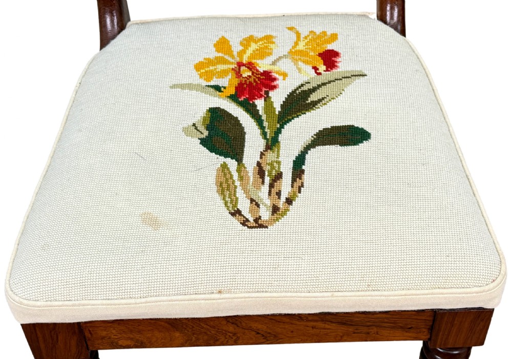 A set of ten Regency rosewood dining chairs featuring carved crest rails, scrolled mid-bars, turned front legs, and cream embroidered needlepoint seats. View X