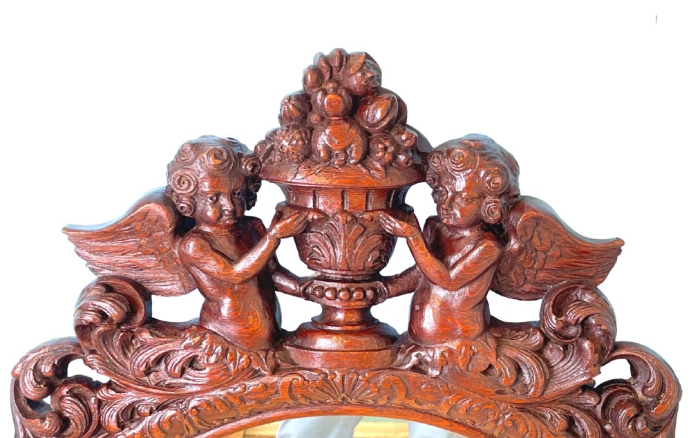 An oval wall mirror framed in carved dark oak, topped with a cresting featuring two cherubs flanking a central urn. View II