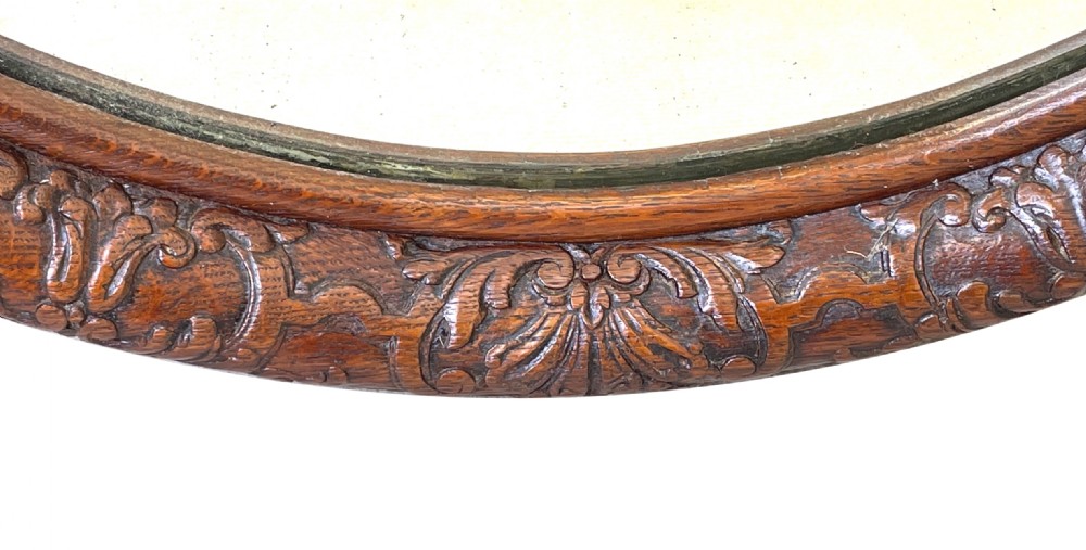 An oval wall mirror framed in carved dark oak, topped with a cresting featuring two cherubs flanking a central urn. View V