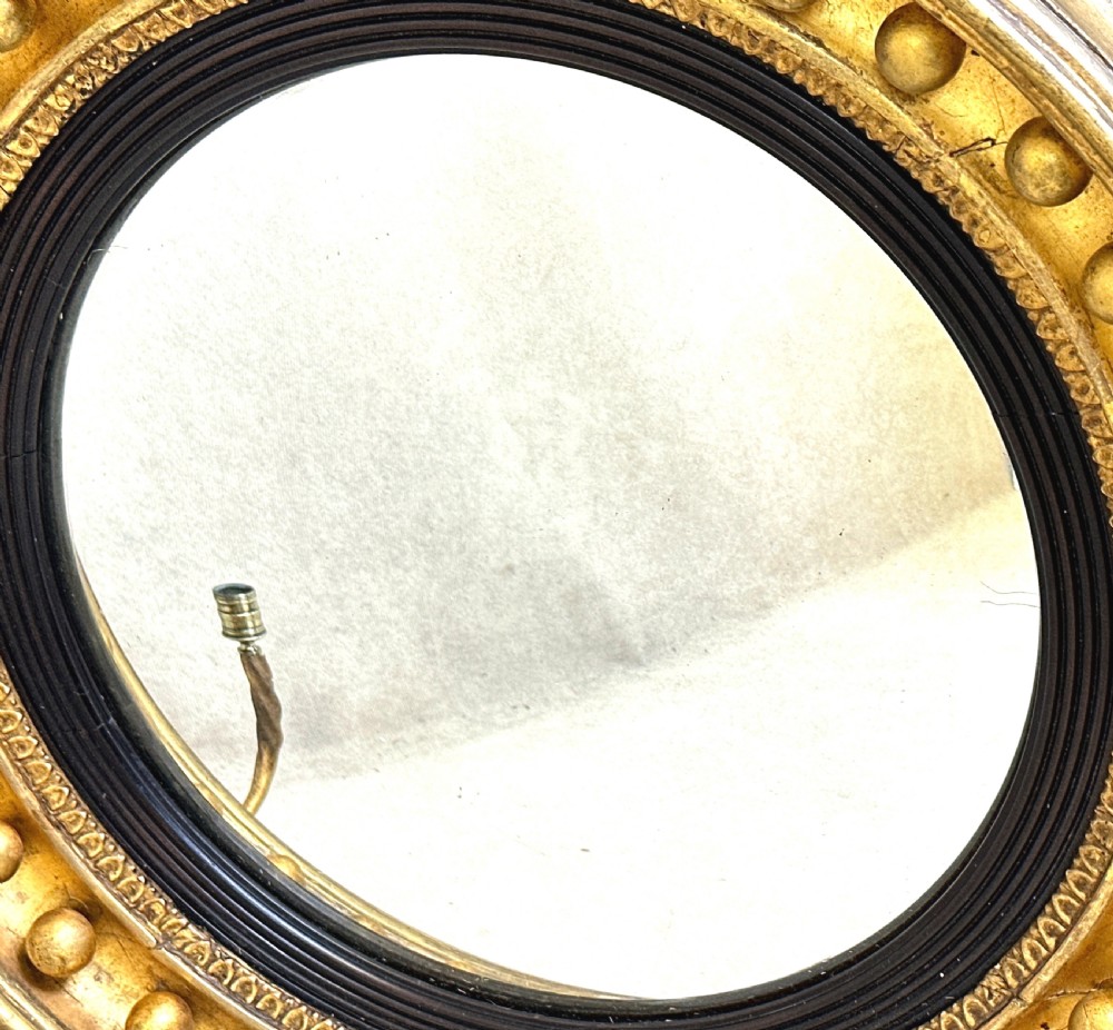 A circular Regency giltwood convex mirror featuring a carved dolphin crest, ball moulding, ebonised inner rim, lower acanthus apron, and twin candle sconces. View VII