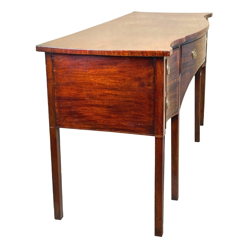 A Georgian mahogany serpentine-front sideboard featuring a polished wood top, central arched drawer, flanking drawers with brass rosette handles, and tapered square legs. View X