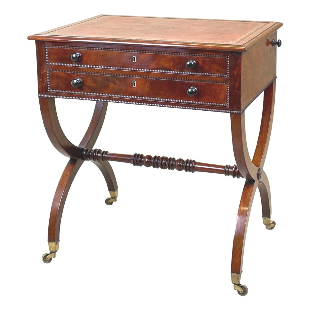 A Regency mahogany writing table featuring a brown leather inset top, two drawers with wooden knobs, curved X-frame legs with a turned stretcher, and brass castors.