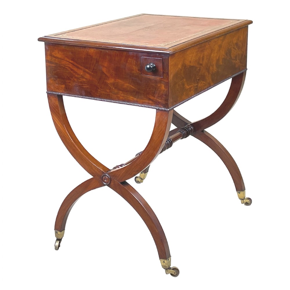 A Regency mahogany writing table featuring a brown leather inset top, two drawers with wooden knobs, curved X-frame legs with a turned stretcher, and brass castors. View II