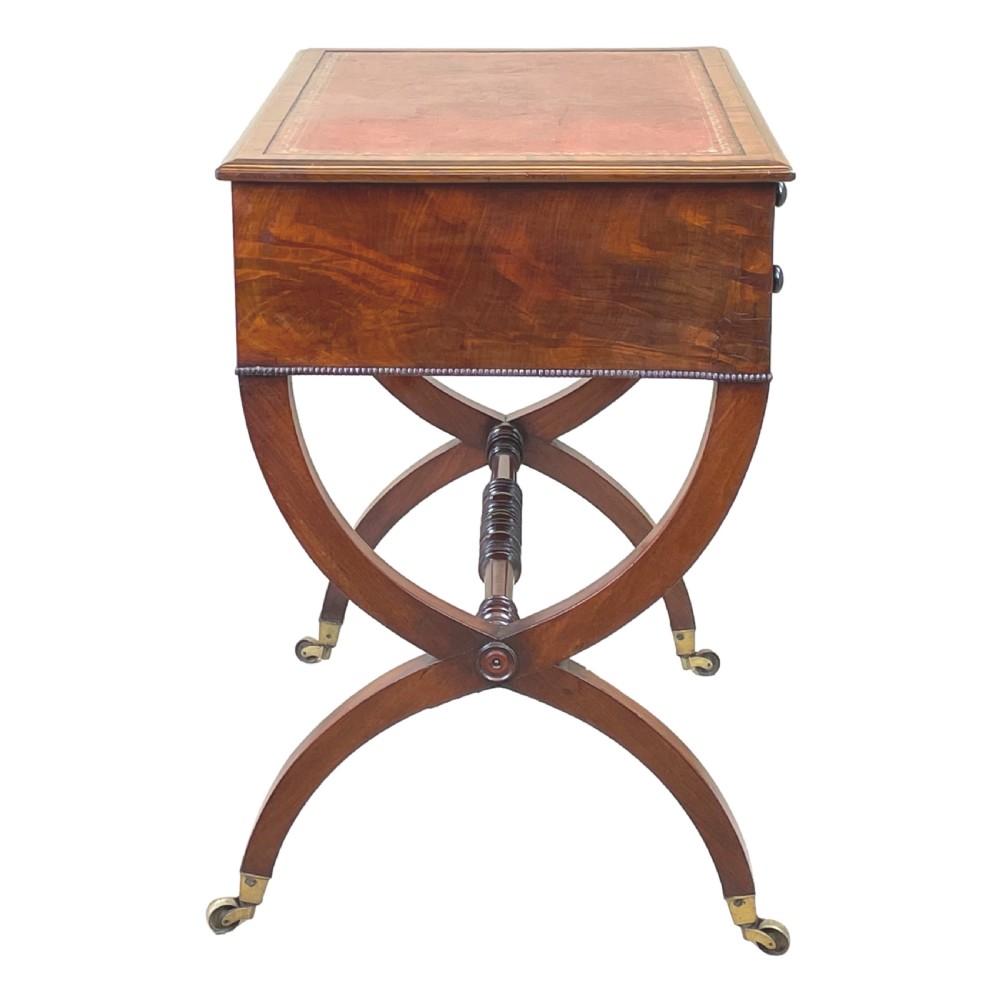 A Regency mahogany writing table featuring a brown leather inset top, two drawers with wooden knobs, curved X-frame legs with a turned stretcher, and brass castors. View III