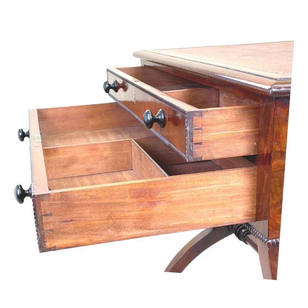A Regency mahogany writing table featuring a brown leather inset top, two drawers with wooden knobs, curved X-frame legs with a turned stretcher, and brass castors. View IV