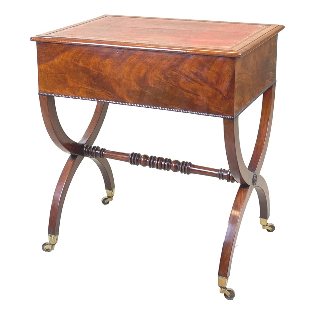 A Regency mahogany writing table featuring a brown leather inset top, two drawers with wooden knobs, curved X-frame legs with a turned stretcher, and brass castors. View VI