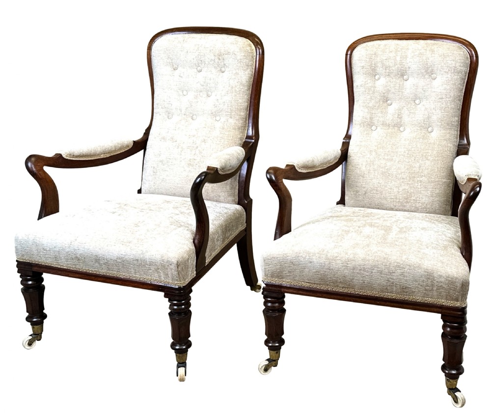 A pair of 19th-century rosewood library armchairs featuring textured beige button-backed upholstery, padded armrests, curved timber supports, turned front legs, and brass castors.