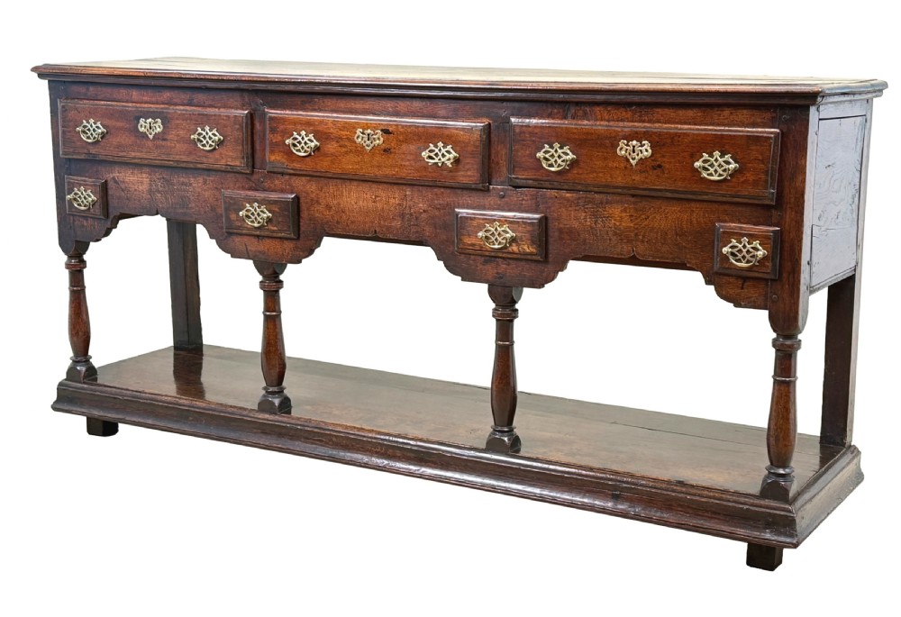An 18th-century dark oak dresser base featuring multiple drawers with pierced brass handles, turned front legs, and a lower pot board shelf.