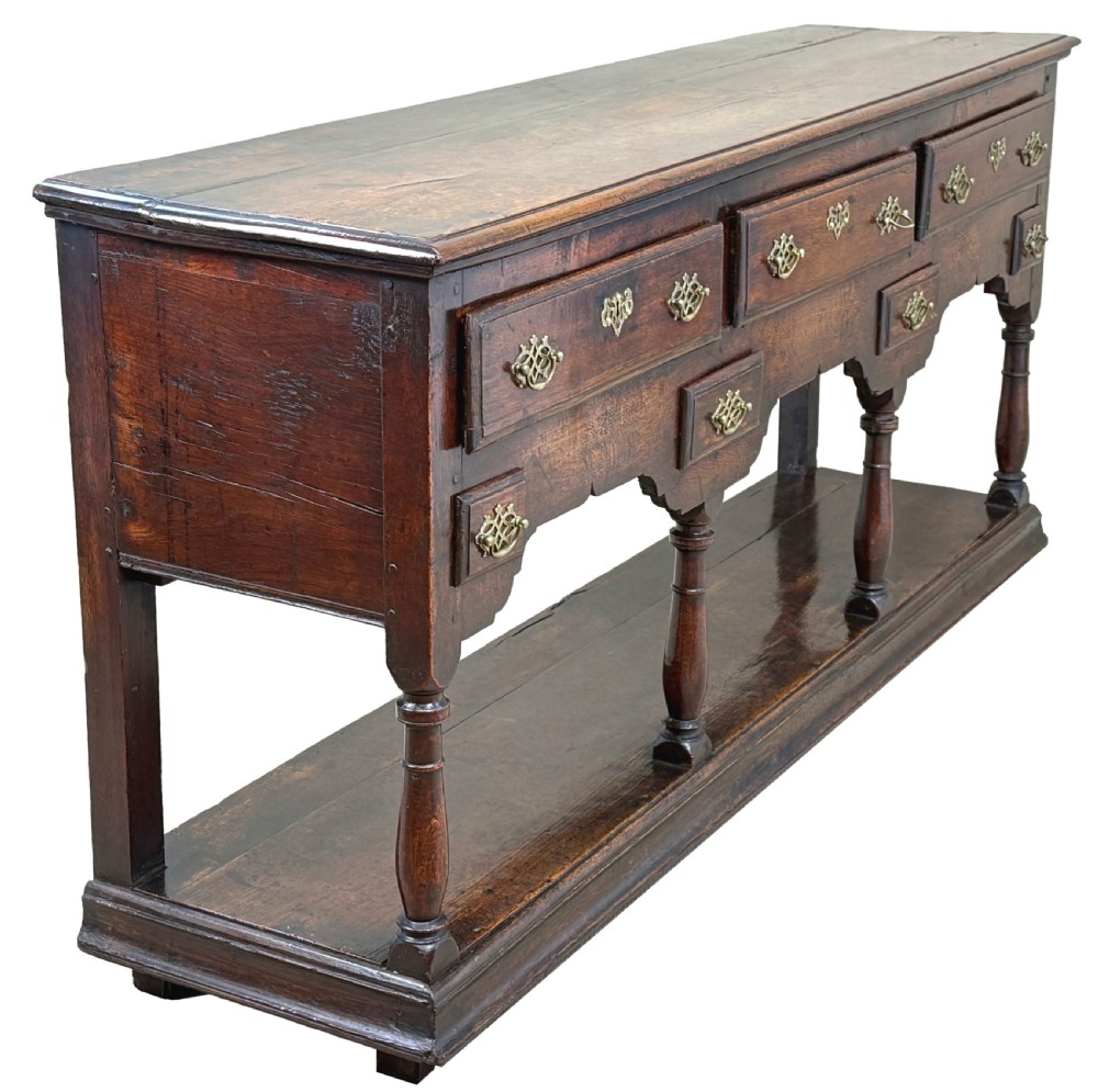 An 18th-century dark oak dresser base featuring multiple drawers with pierced brass handles, turned front legs, and a lower pot board shelf. View VI