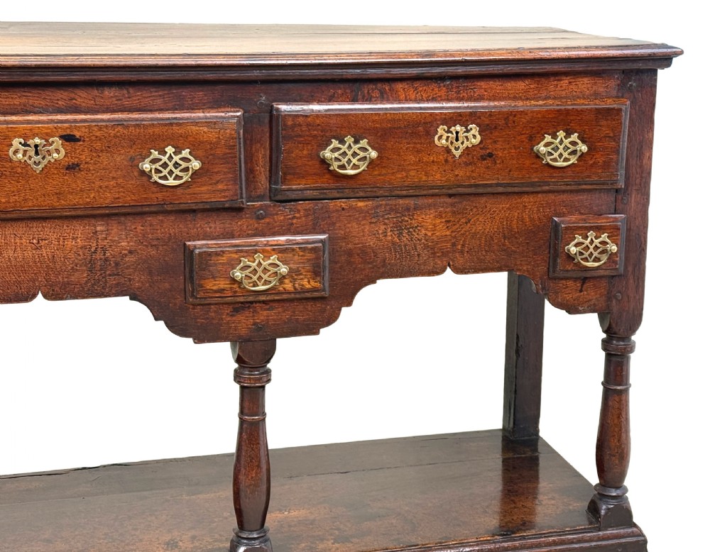 An 18th-century dark oak dresser base featuring multiple drawers with pierced brass handles, turned front legs, and a lower pot board shelf. View VII