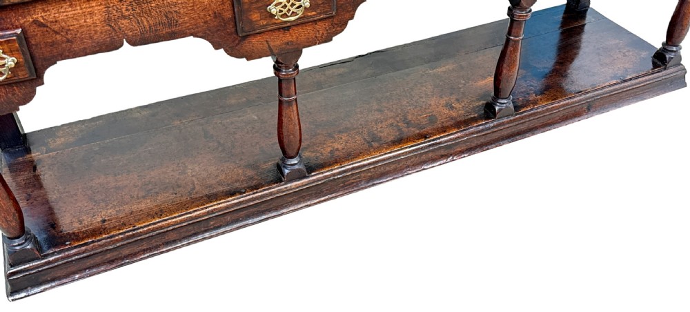An 18th-century dark oak dresser base featuring multiple drawers with pierced brass handles, turned front legs, and a lower pot board shelf. View X