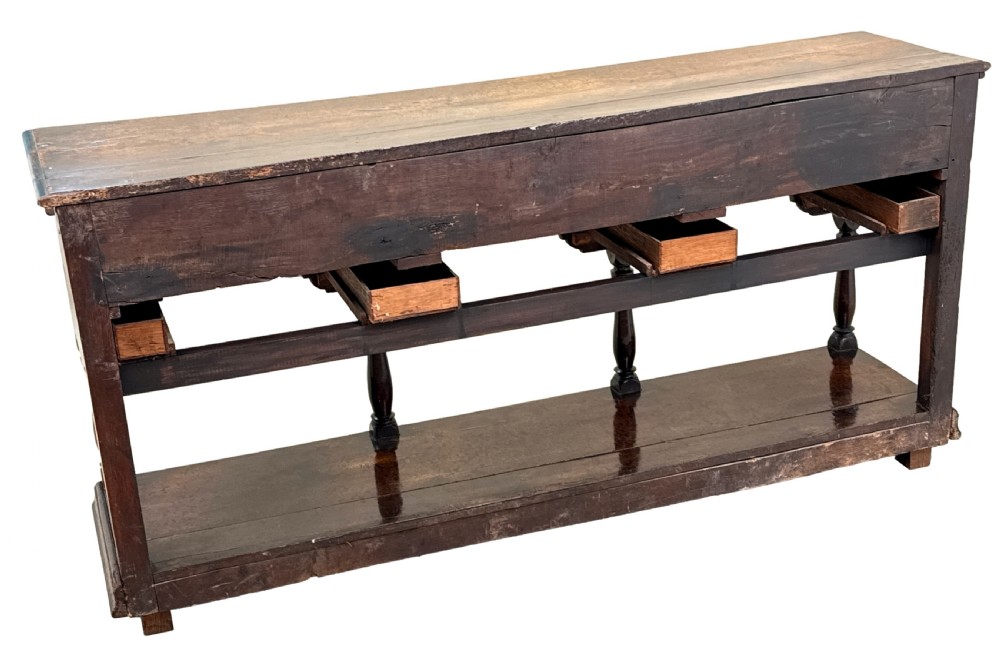 An 18th-century dark oak dresser base featuring multiple drawers with pierced brass handles, turned front legs, and a lower pot board shelf. View XII
