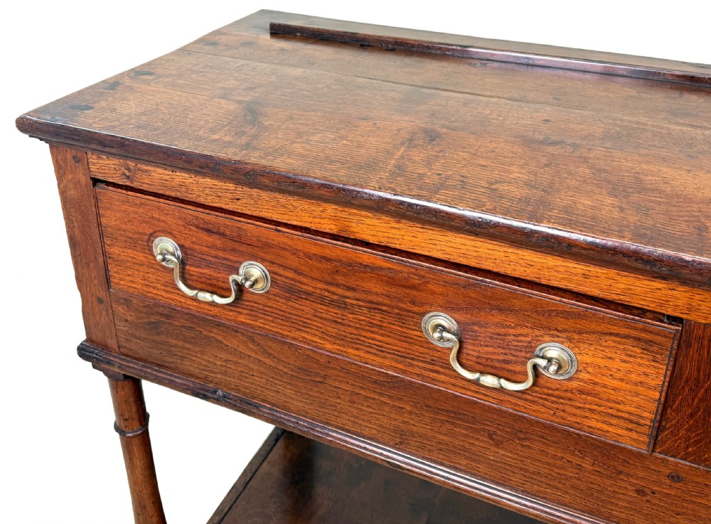 A late 18th-century Georgian oak dresser base featuring two drawers with brass swan-neck handles, turned front legs, and a lower pot board shelf. View III