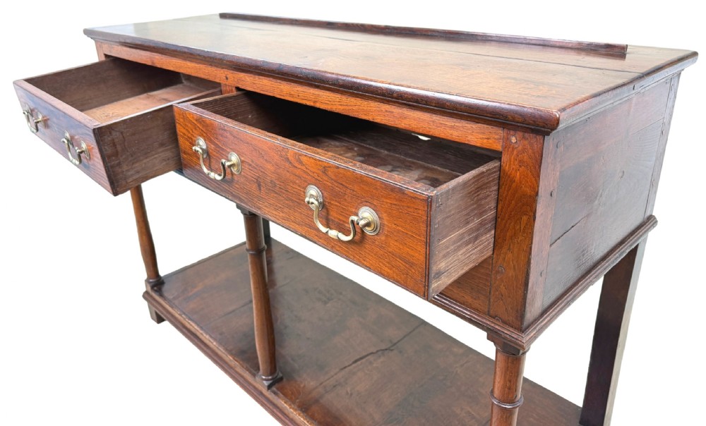 A late 18th-century Georgian oak dresser base featuring two drawers with brass swan-neck handles, turned front legs, and a lower pot board shelf. View IV