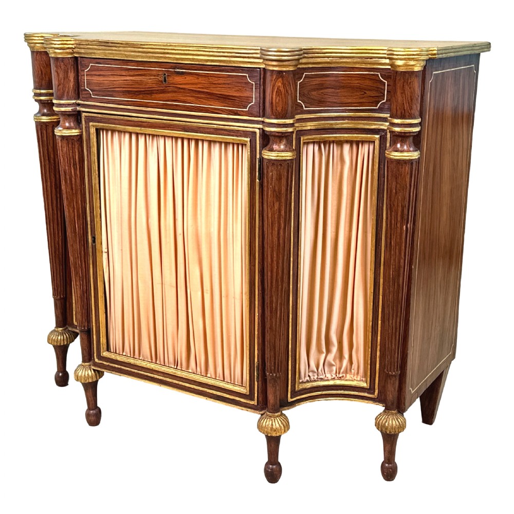An antique Regency style breakfront side cabinet featuring simulated rosewood finishing, gold leaf gilded columns, and doors set with pleated pink fabric.