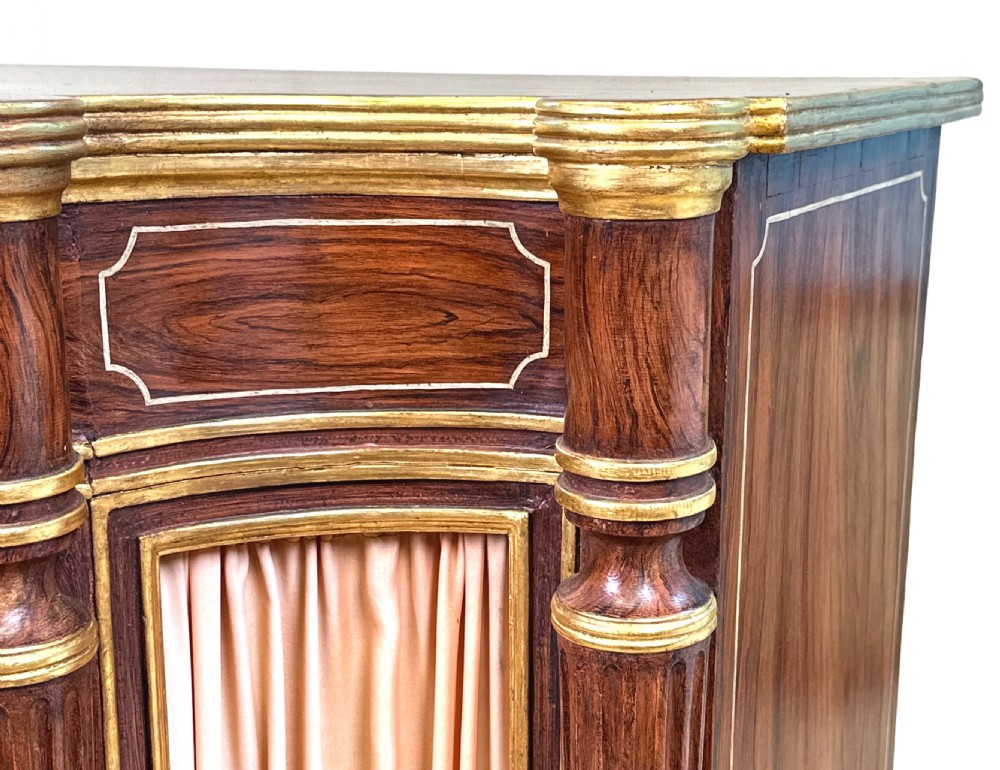 An antique Regency style breakfront side cabinet featuring simulated rosewood finishing, gold leaf gilded columns, and doors set with pleated pink fabric. View VI