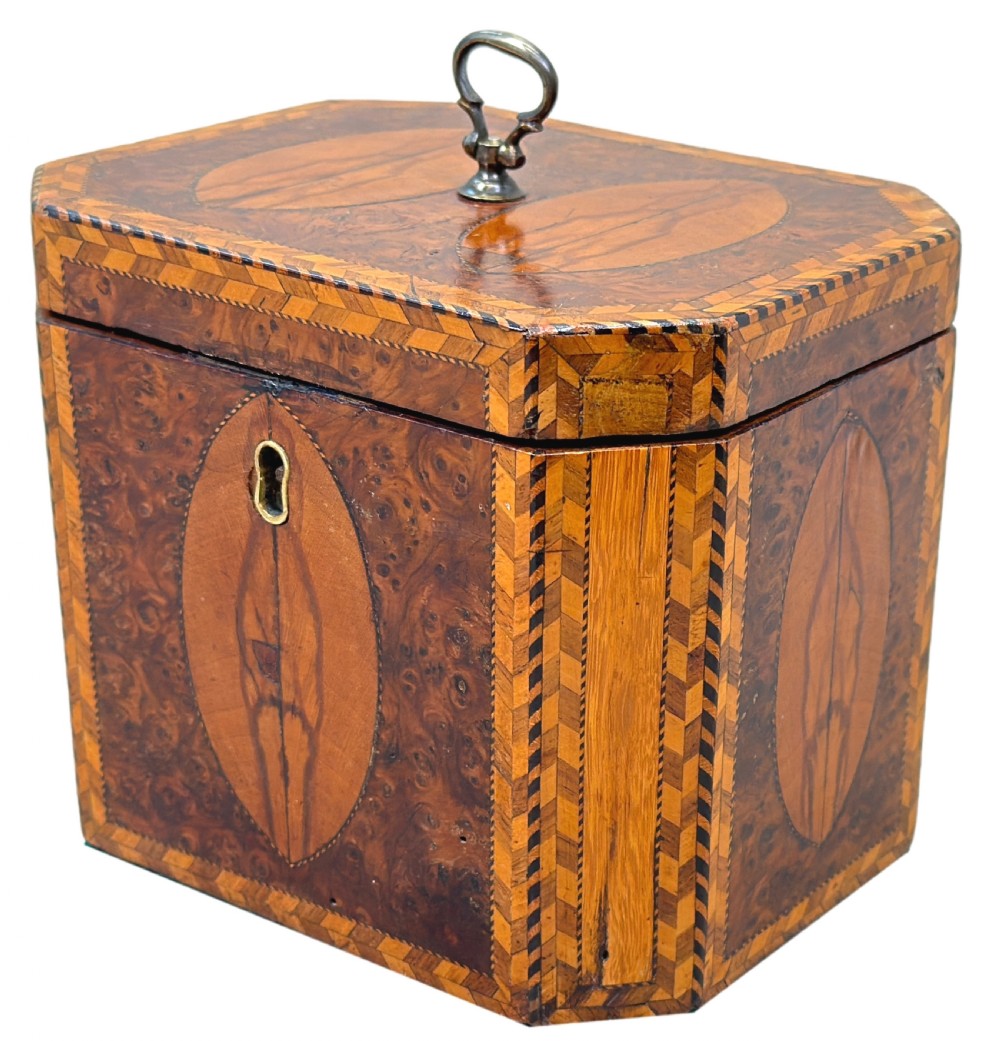 An 18th-century octagonal Georgian tea caddy crafted from burr yew wood with chevron stringing, oval marquetry panels, a brass top handle, and a brass keyhole escutcheon.