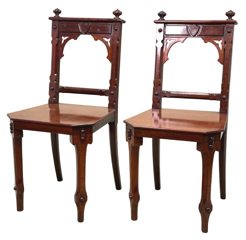 A pair of Victorian mahogany hall chairs featuring carved shield backrests, open architectural arches, turned finials, and solid wooden seats.
