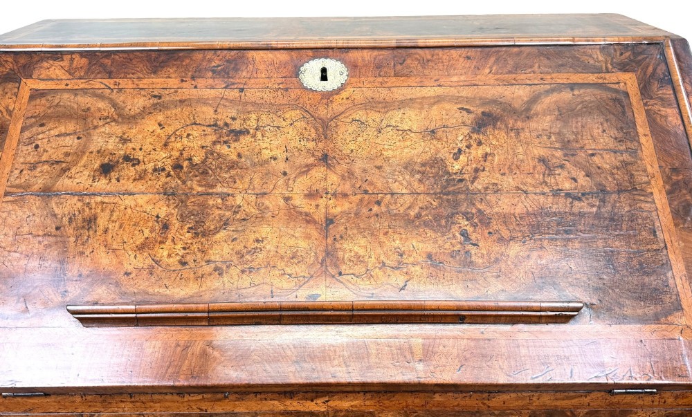 An early 18th-century Queen Anne walnut bureau featuring a slanted fall-front writing flap, brass escutcheons with drop handles, and turned bun feet. View X