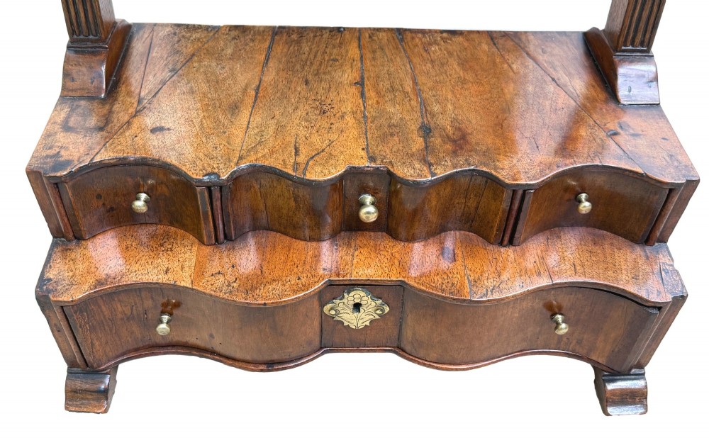 A Queen Anne early 18th-century walnut dressing table mirror featuring an arched swing frame resting above a stepped serpentine base with small drawers and brass hardware. View VI