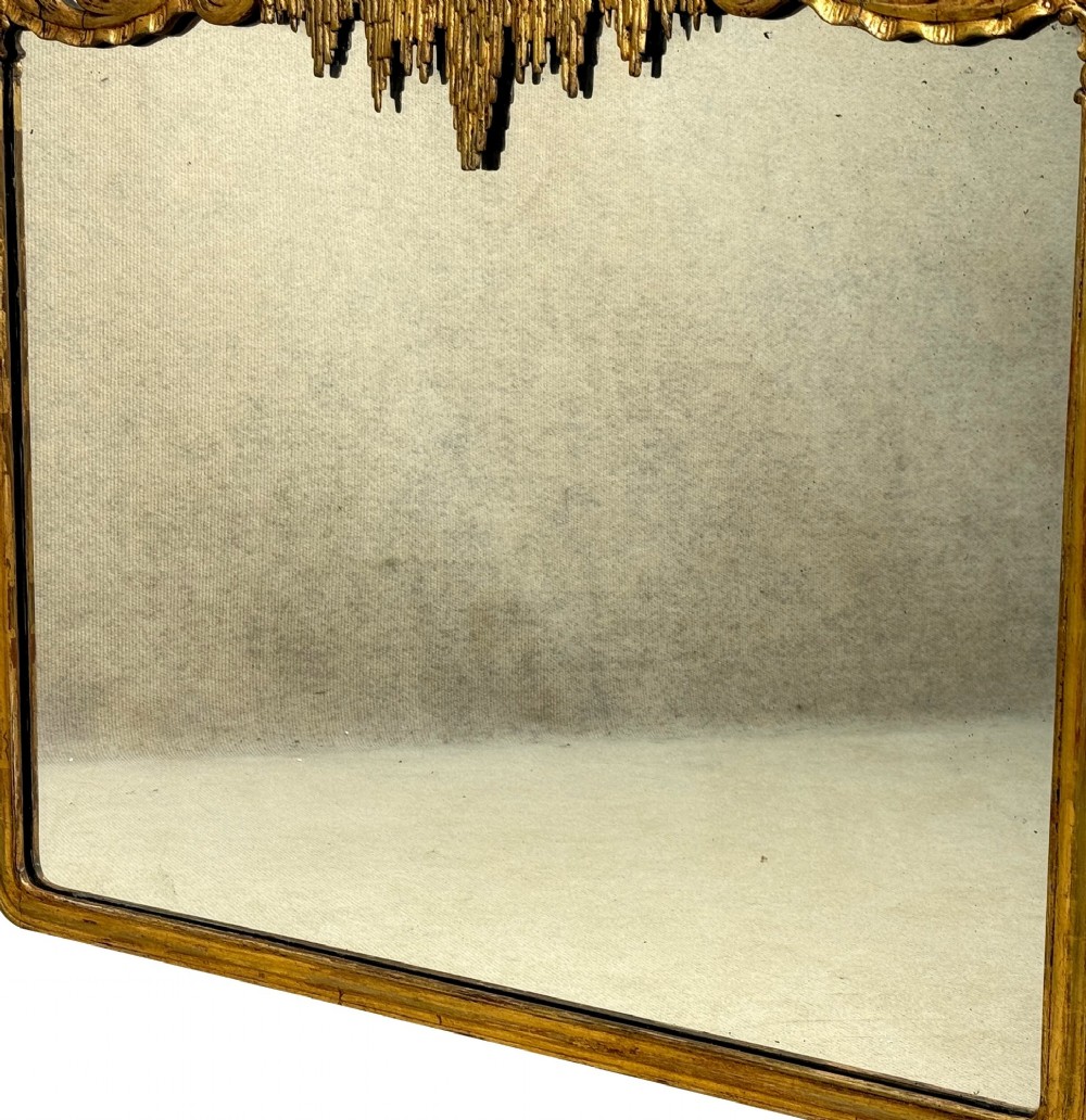 A late 19th-century giltwood wall mirror featuring a carved gesso cresting with scrollwork and dripping fringe details above a rectangular glass panel. View V