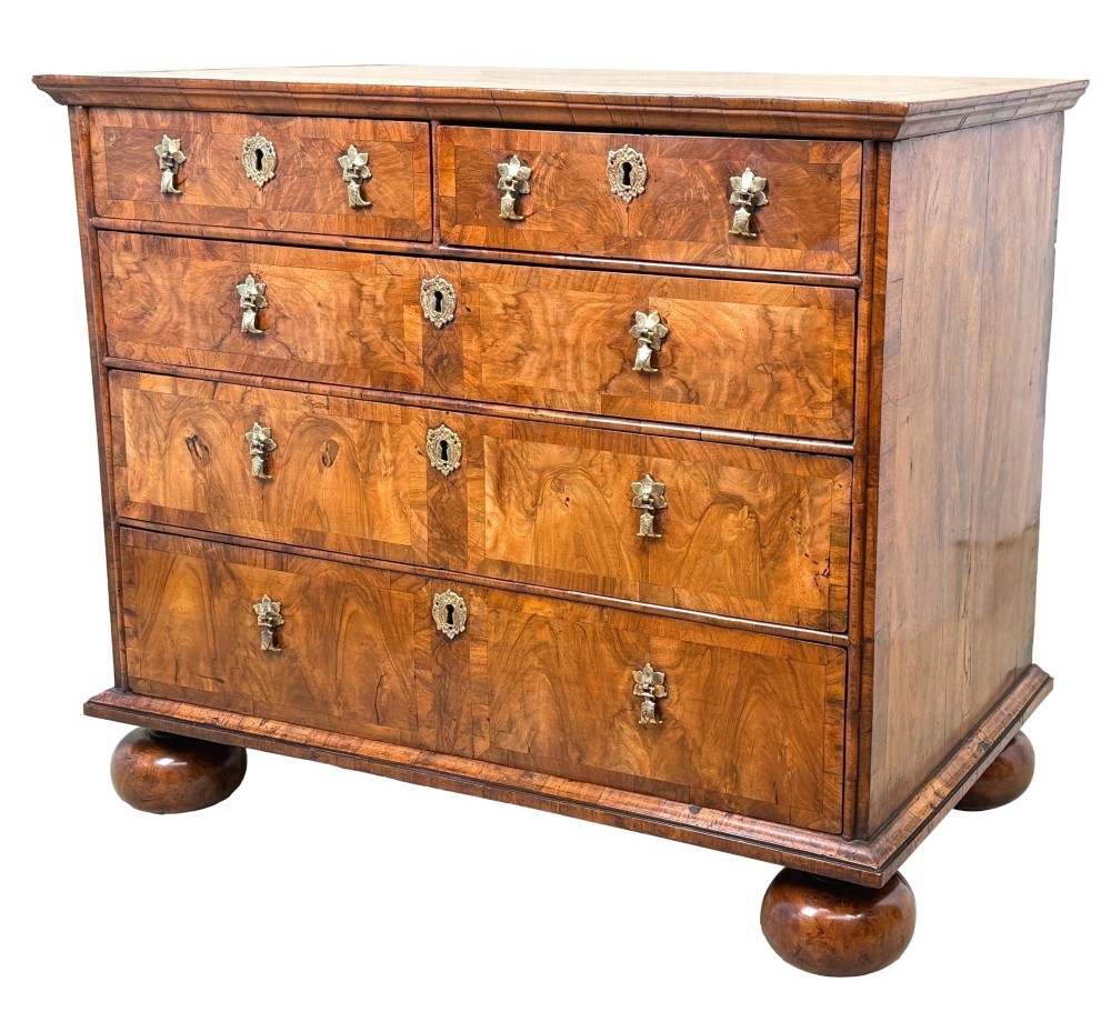 An early 18th-century Queen Anne walnut chest of drawers featuring figured grain veneers, brass drop handles, and turned bun feet.