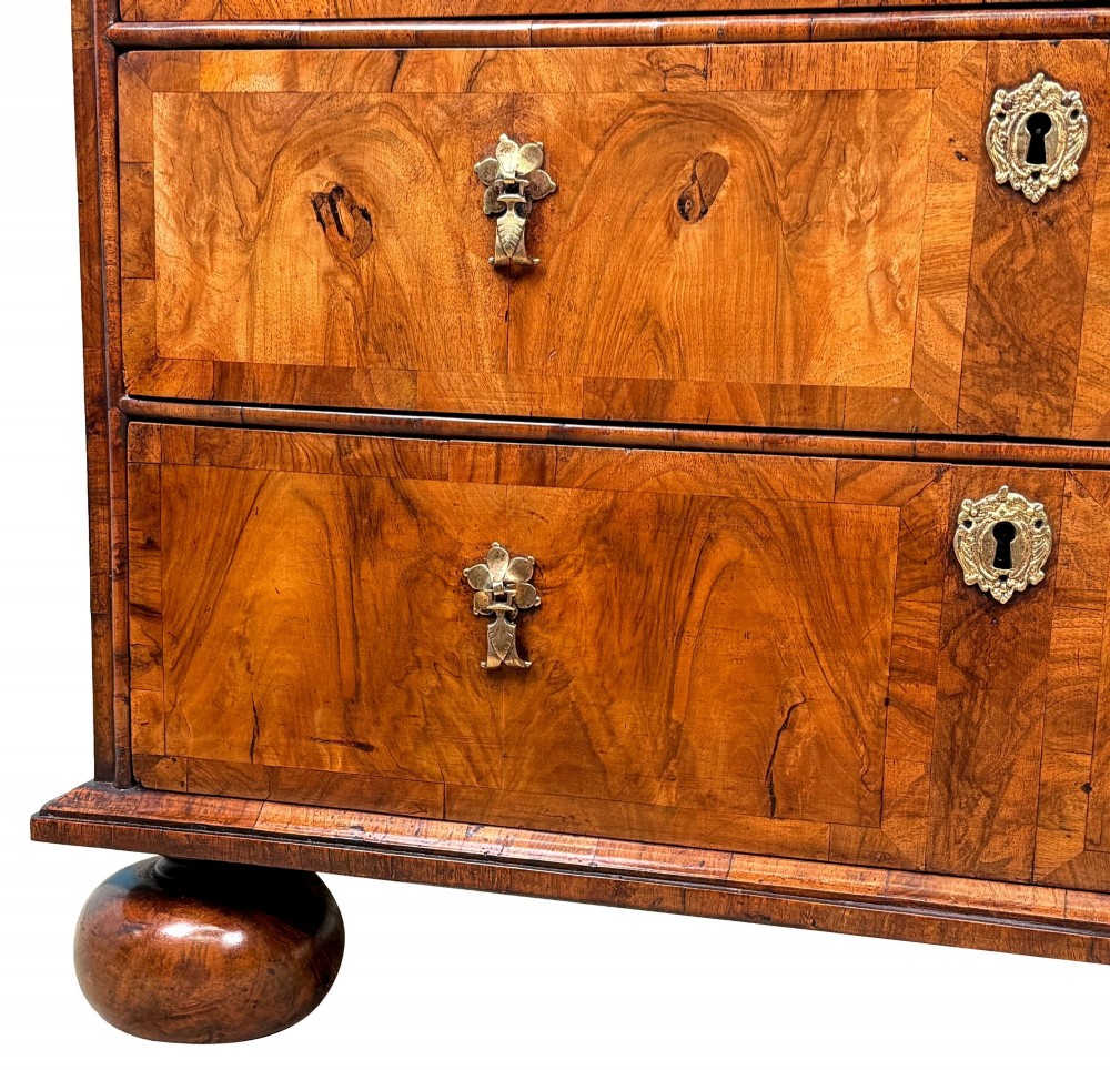 An early 18th-century Queen Anne walnut chest of drawers featuring figured grain veneers, brass drop handles, and turned bun feet. View VII