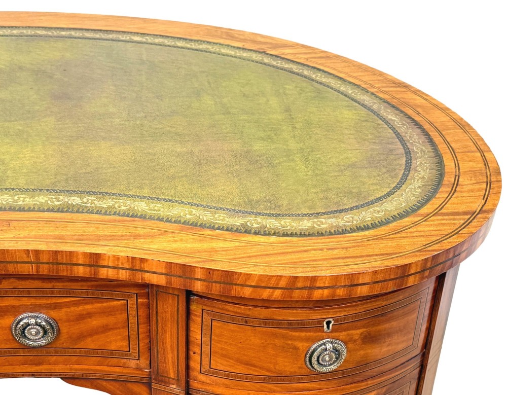 A 19th-century kidney-shaped satinwood writing desk featuring an inset green leather top, nine inlaid drawers with brass ring handles, and tapered square legs on castors. View VIII