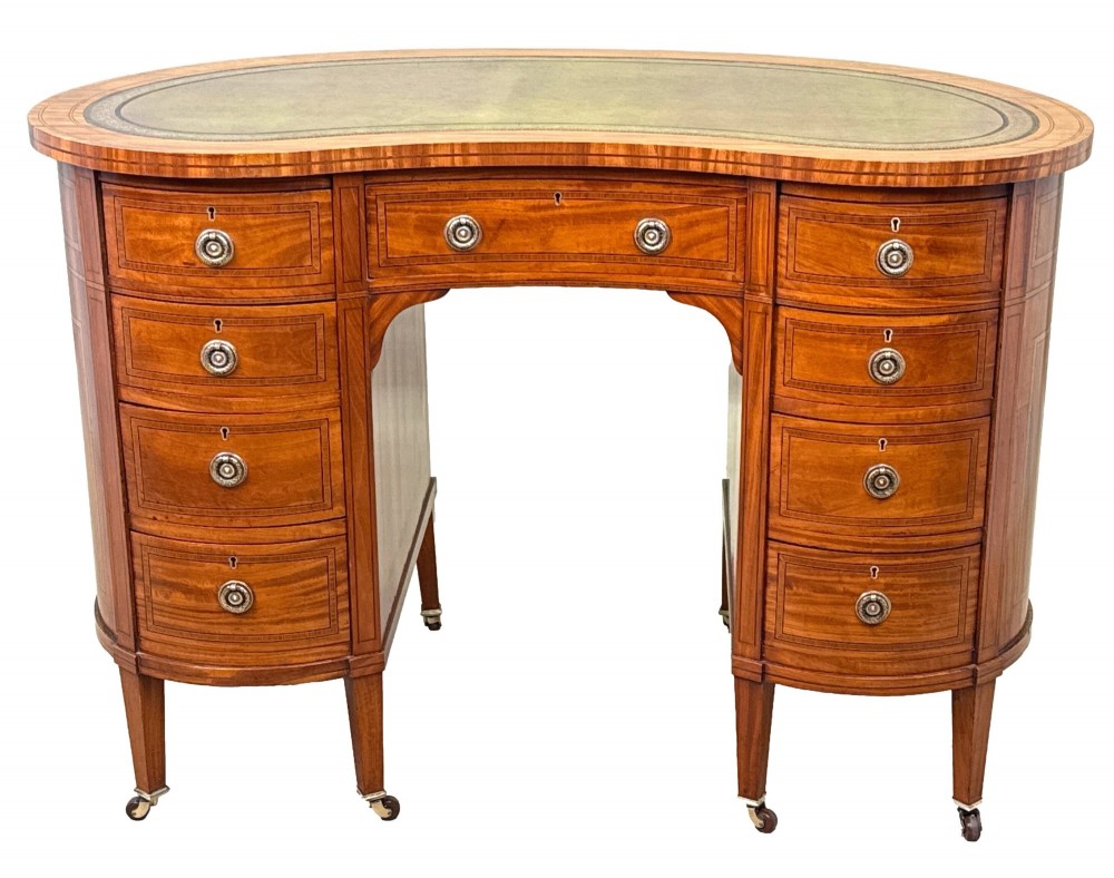 A 19th-century kidney-shaped satinwood writing desk featuring an inset green leather top, nine inlaid drawers with brass ring handles, and tapered square legs on castors. View X