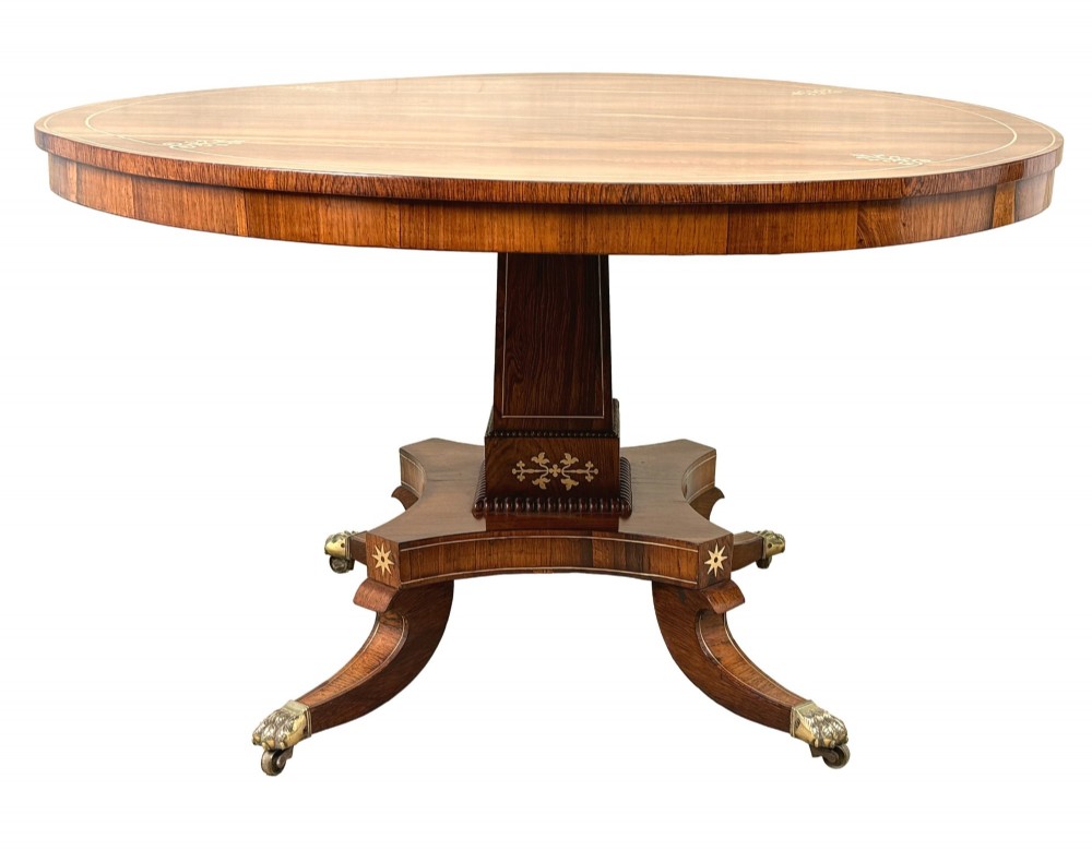 A circular Regency rosewood centre table featuring brass inlay motifs around the rim, a central pedestal support, and a quadform base with splayed legs ending in brass paw castors.
