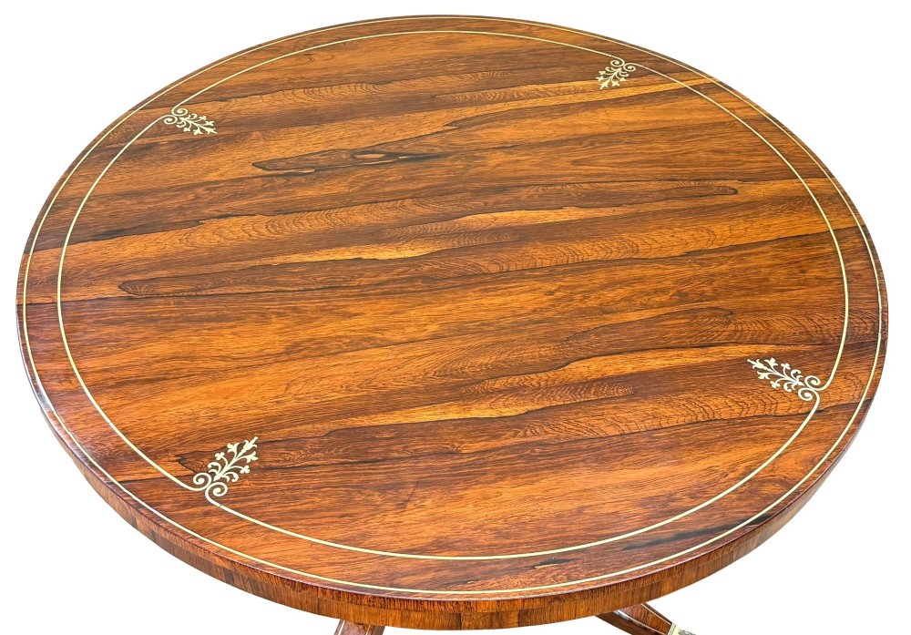A circular Regency rosewood centre table featuring brass inlay motifs around the rim, a central pedestal support, and a quadform base with splayed legs ending in brass paw castors. View II