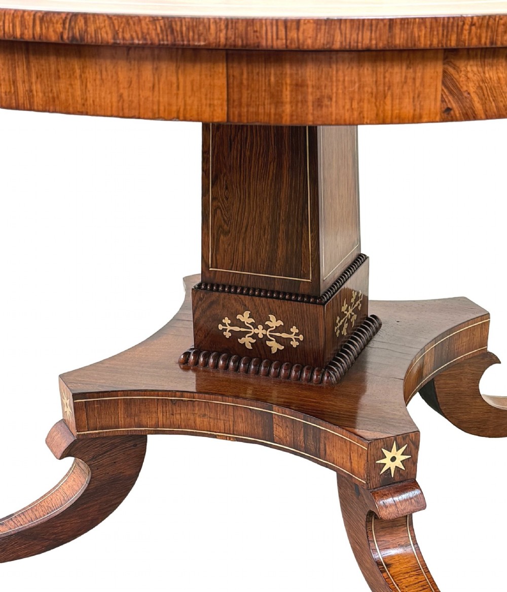 A circular Regency rosewood centre table featuring brass inlay motifs around the rim, a central pedestal support, and a quadform base with splayed legs ending in brass paw castors. View III