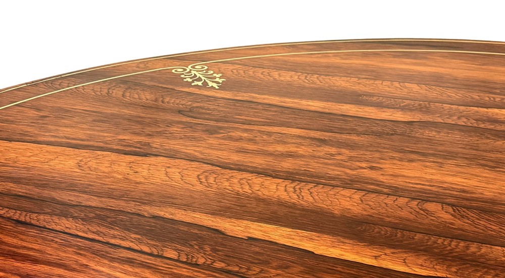 A circular Regency rosewood centre table featuring brass inlay motifs around the rim, a central pedestal support, and a quadform base with splayed legs ending in brass paw castors. View V