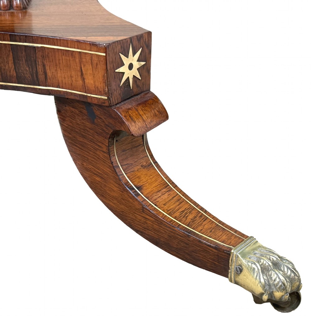 A circular Regency rosewood centre table featuring brass inlay motifs around the rim, a central pedestal support, and a quadform base with splayed legs ending in brass paw castors. View VII